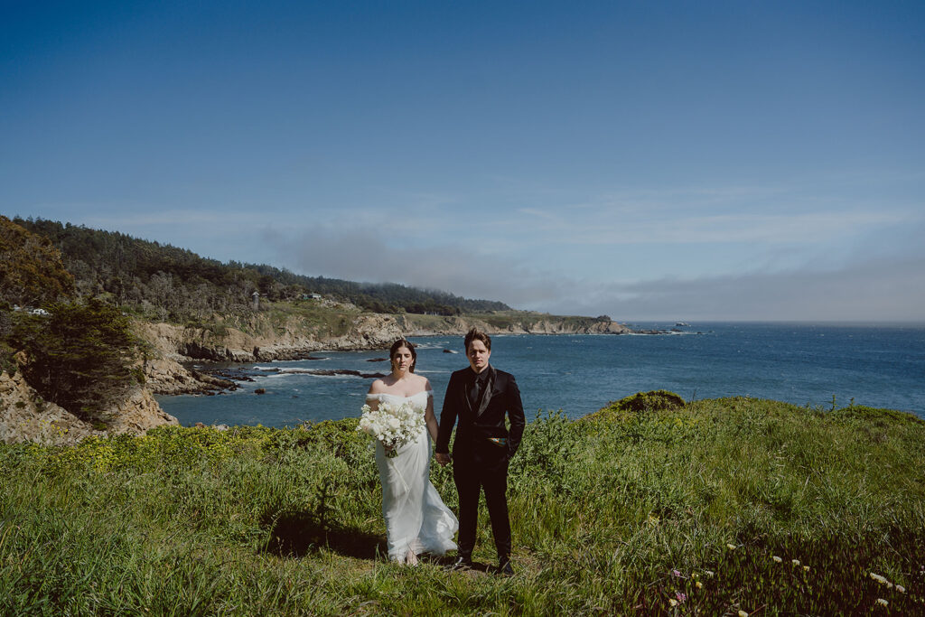 A bride and groom stand together on a grassy cliff overlooking the ocean, with a rocky coastline and clear blue sky in the background, perfectly capturing the magic crafted by their coastal wedding planner.