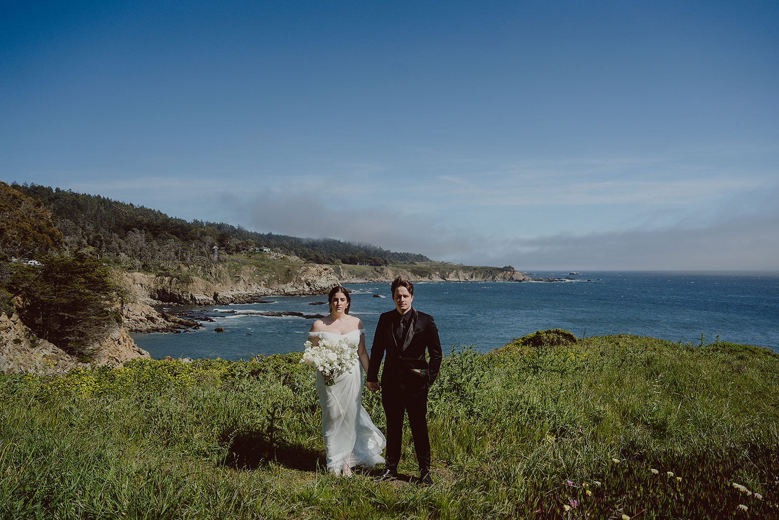 A bride and groom stand together on a grassy cliff overlooking the ocean, with a rocky coastline and clear blue sky in the background, perfectly capturing the magic crafted by their coastal wedding planner.