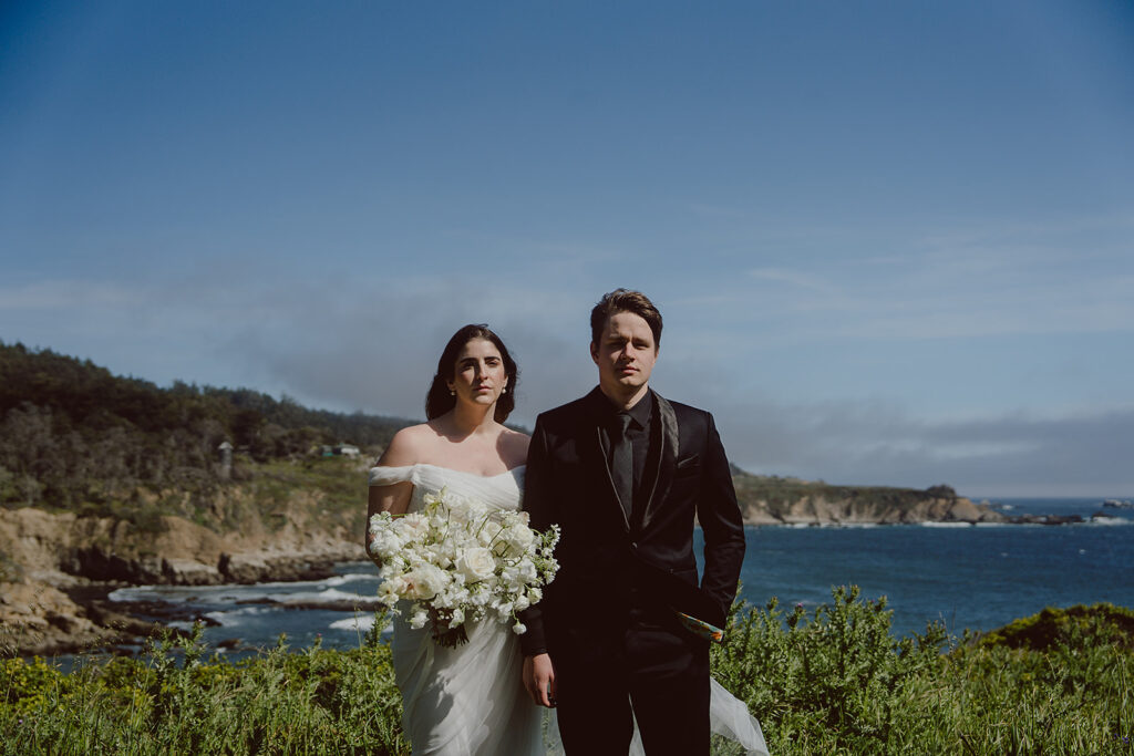 A bride and groom stand outdoors with a scenic coastal landscape in the background, including cliffs, ocean, and clear sky. The bride holds a bouquet of white flowers.