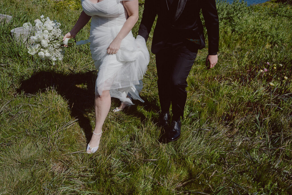 A couple strolls through a grassy field, the woman in a white dress holding a bouquet and the man in a dark suit, guided by their expert coastal wedding planner.