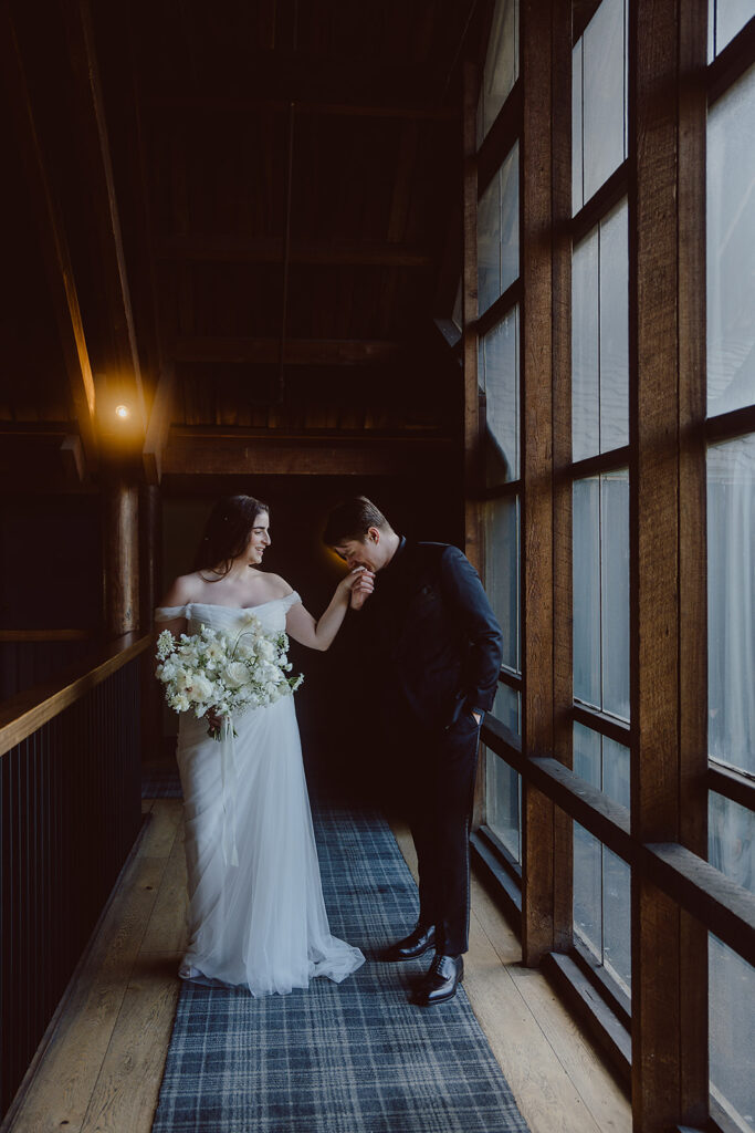 A bride in a white dress holds a bouquet while a man in a dark suit kisses her hand in a dimly lit hallway with large windows.