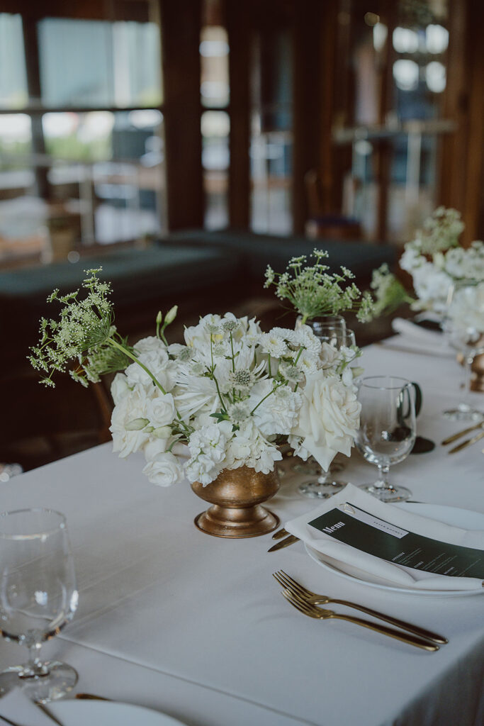 Elegant table setting with white floral centerpieces, clear glassware, and white plates arranged on a white tablecloth. Gold utensils are placed neatly beside each setting.