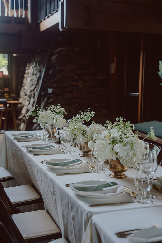 A dining table set for six with white tablecloth, white flowers in gold vases, neatly arranged napkins, plates, cutlery, and glasses.