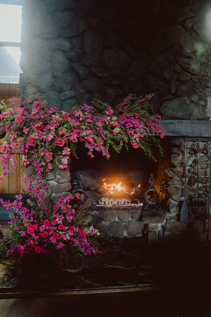 A stone fireplace with a lit fire, adorned with vibrant pink flowers and greenery on the mantel and floor, evokes the serene charm of a coastal wedding planner's dream setting.