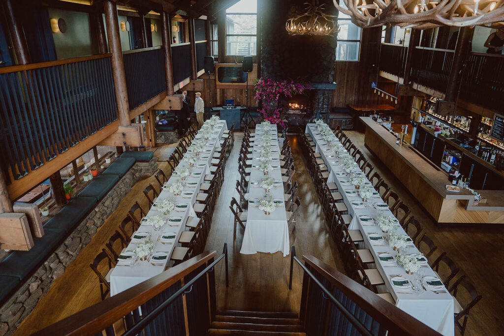 A long dining hall with two parallel rows of tables set for a banquet. The room features wooden beams, a stone fireplace, and hanging chandeliers.