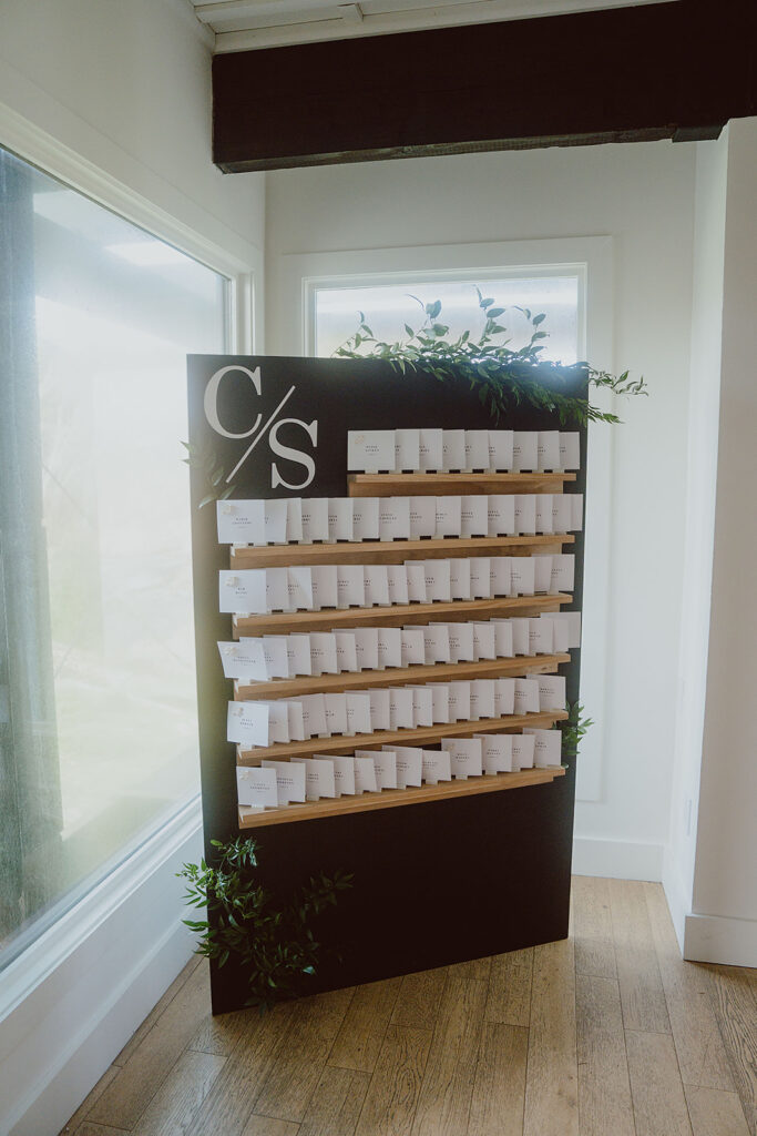 A black display board with rows of envelopes arranged neatly, adorned with green foliage, placed in a room with wooden floors and large windows.
