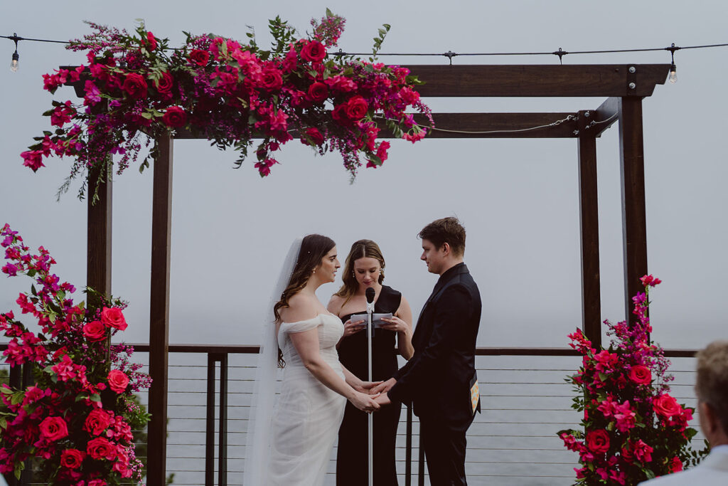 A bride and groom hold hands under a wooden arch adorned with vibrant pink flowers. An officiant stands in the center, reading from a script.