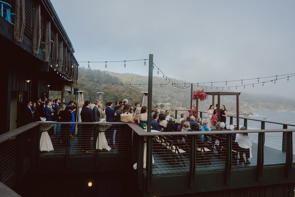 Outdoor wedding ceremony overlooking the ocean, with guests seated and standing on a wooden deck adorned with string lights and floral arrangements.