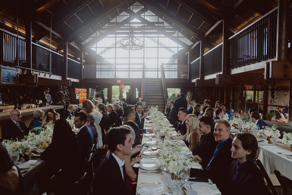 A large group of people seated at long tables in a rustic venue with high ceilings, stairway, and floral decorations.