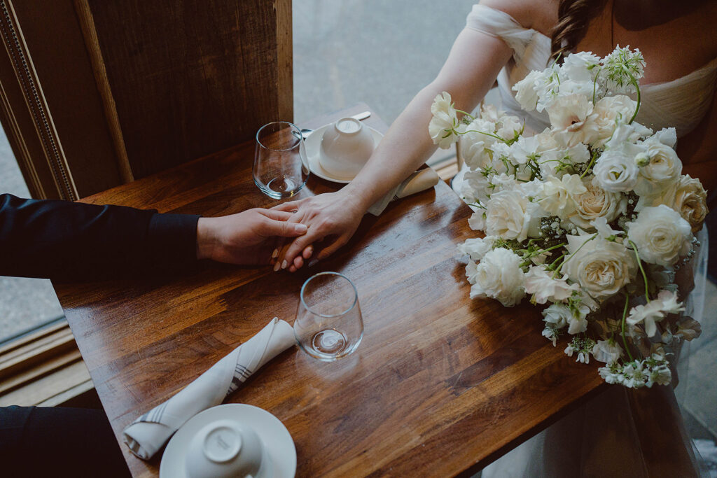 Bride holding a large white flower bouquet, sitting at a wooden table, reaching across to hold hands with someone. Two glasses and napkins are on the table.
