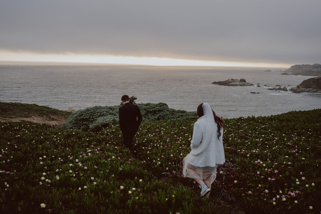A couple walking on a grassy hillside with wildflowers near the ocean under a cloudy sky at sunset.