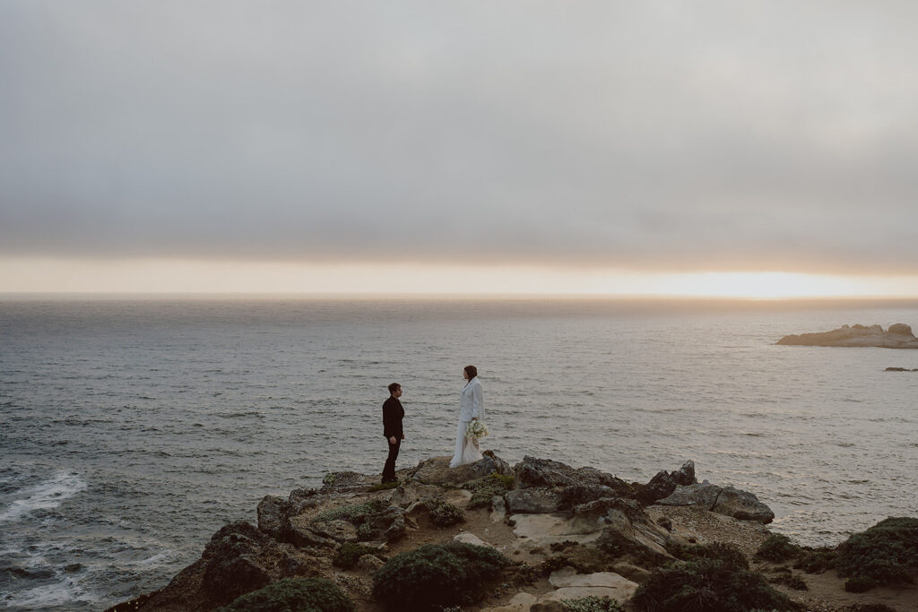 Two people stand on a rocky cliff overlooking the ocean at sunset, with cloudy skies above.