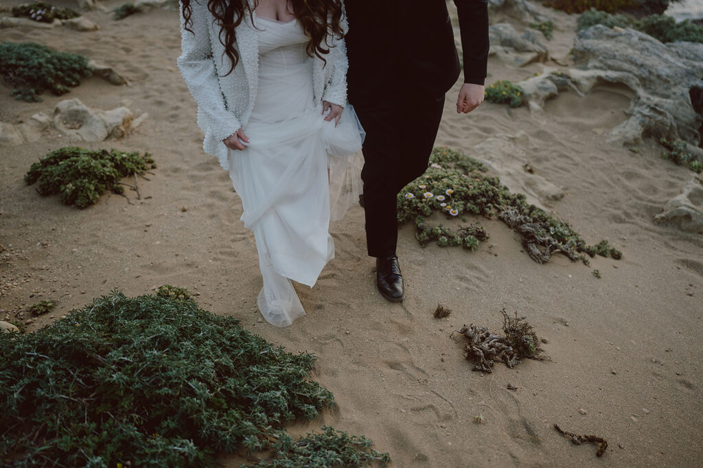 Bride and groom walking side by side on sandy terrain with greenery and rocks. Bride wears a white dress; groom wears a black suit.