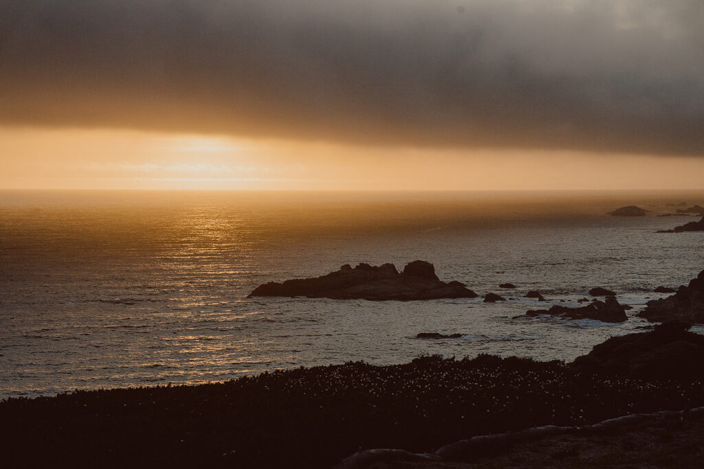 Sunset over a calm ocean with dark clouds above and rocky coastline in the foreground.