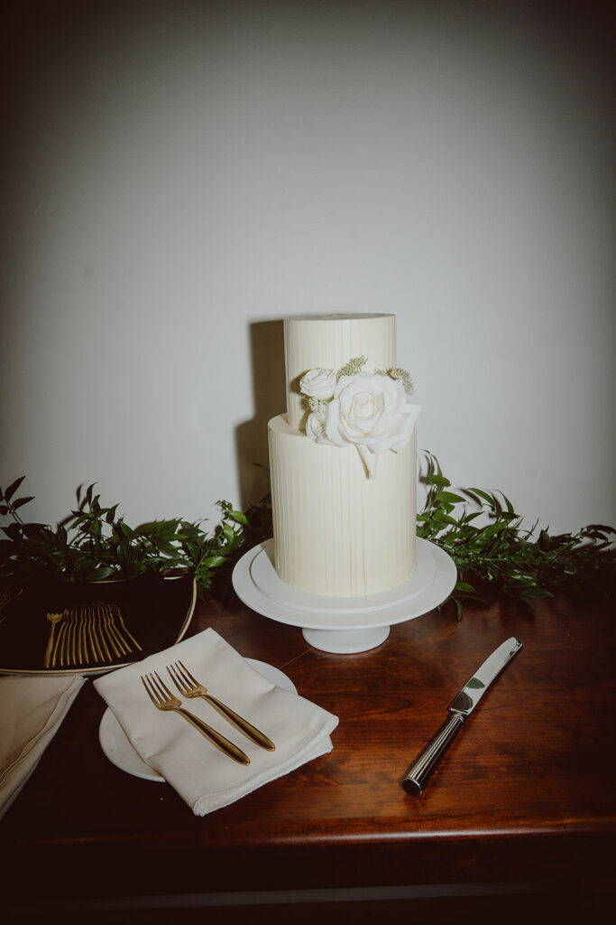 A two-tier white cake with floral decor on a cake stand surrounded by greenery. A knife and a plate with a fork and napkin are on the wooden table next to it.
