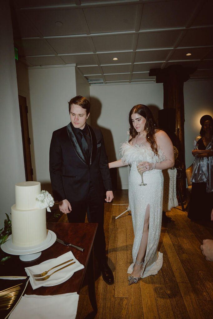 A couple stands near a decorated cake on a wooden table. The woman holds a wine glass and gestures towards the cake. Both are dressed in formal wear.