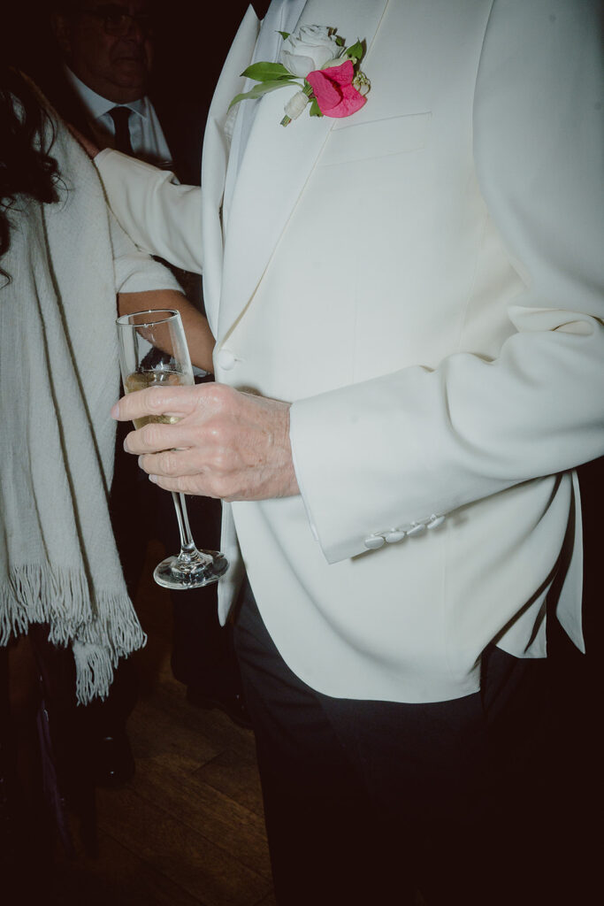 A person in a white suit holding a champagne glass, with a red flower and greenery in the lapel, stands next to another person wearing a shawl.