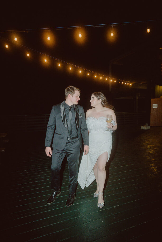 A couple walks outdoors at night under string lights. The man wears a dark suit, and the woman is in a white gown holding a drink.