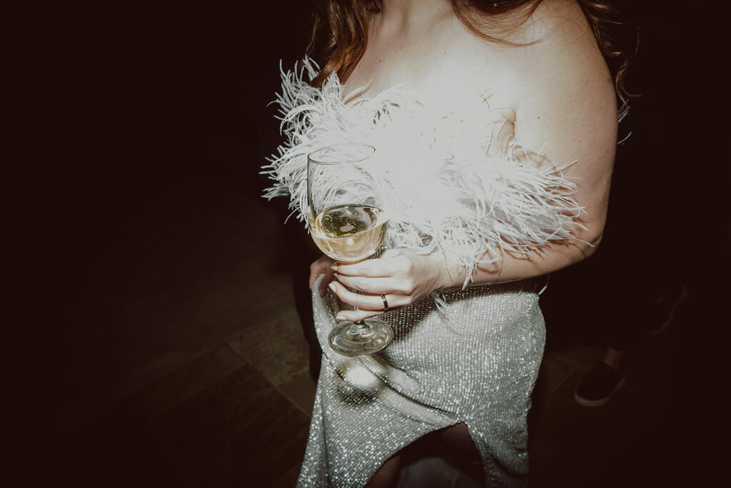Person in a sparkly outfit holding a glass of white wine, with a feathered detail on their top.