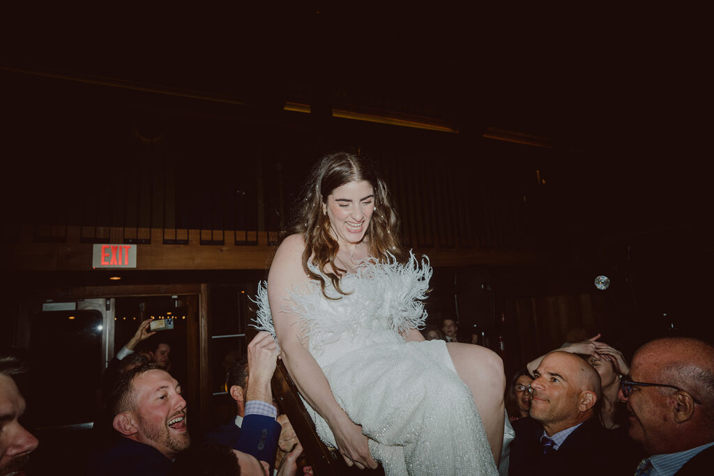 A woman in a white dress is lifted on a chair by a group of people indoors. She is smiling and laughing.