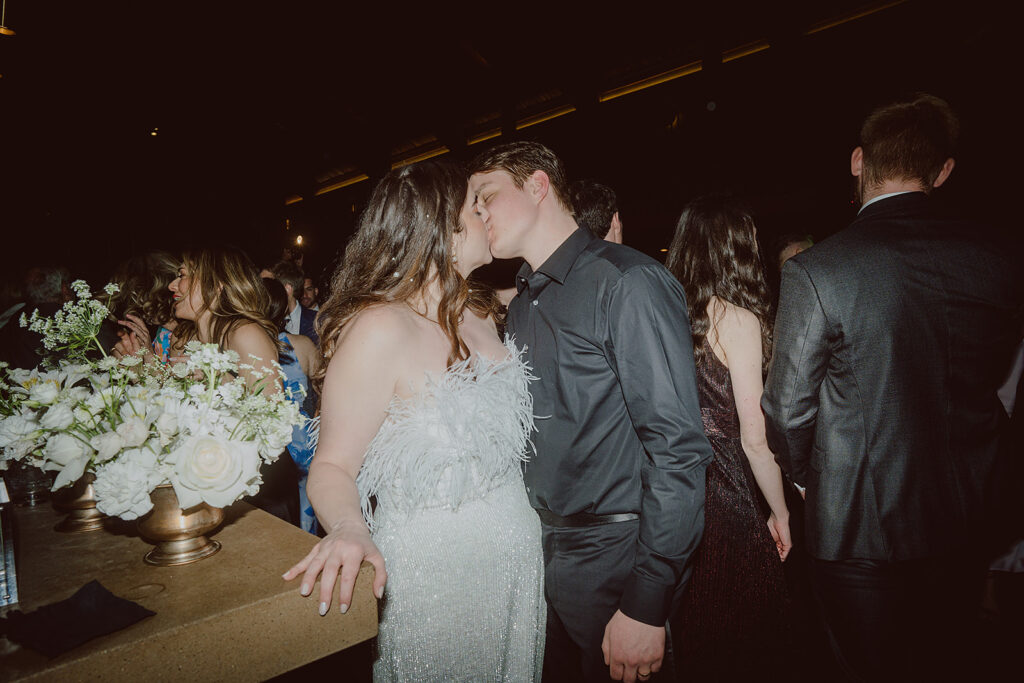 A couple in formal attire shares a kiss at an indoor event, surrounded by other attendees. The woman wears a white, feathered outfit, and the man is in a black shirt. White flowers are on the table nearby.
