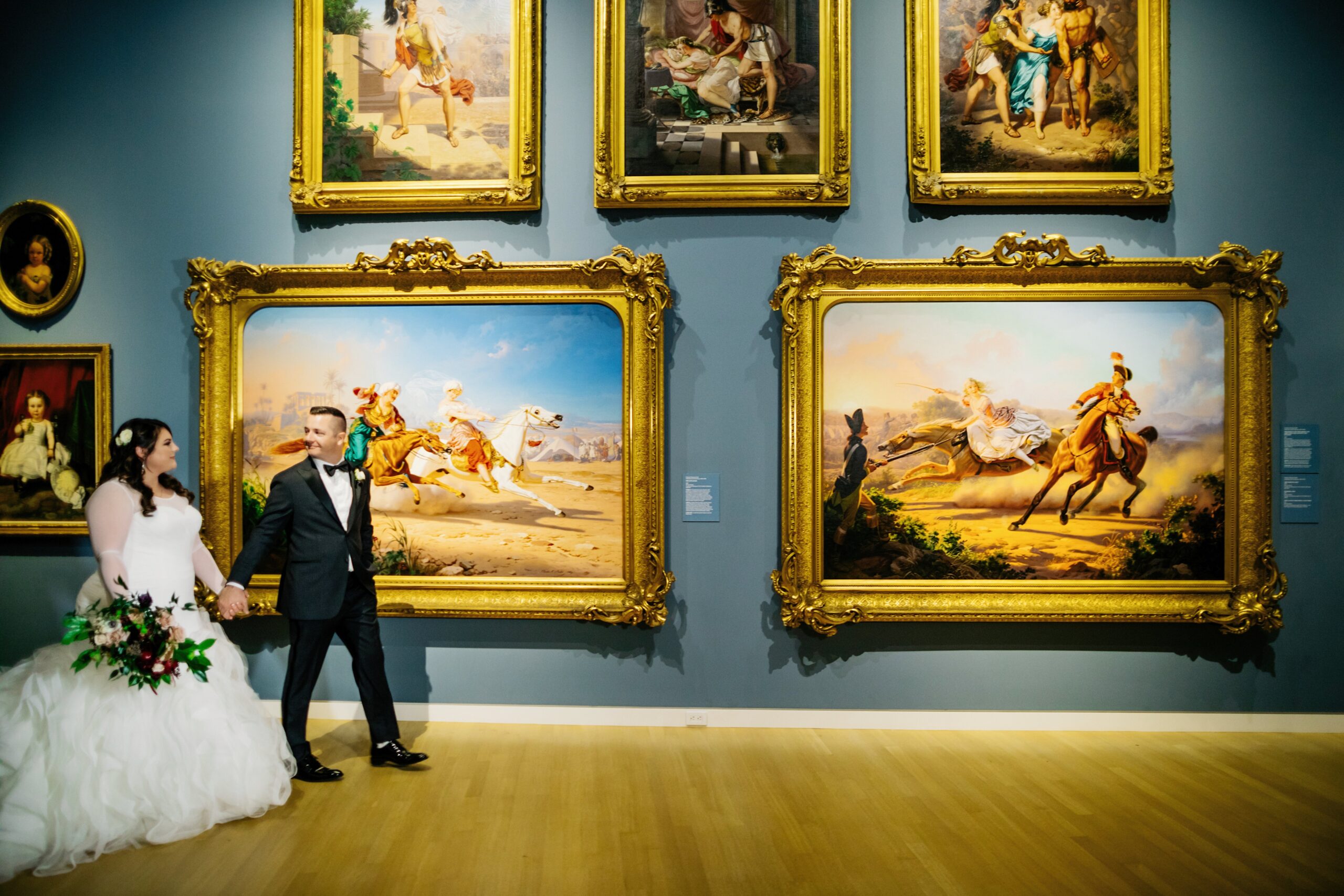 A bride and groom walk hand in hand through an art gallery, surrounded by large, framed paintings on the walls.