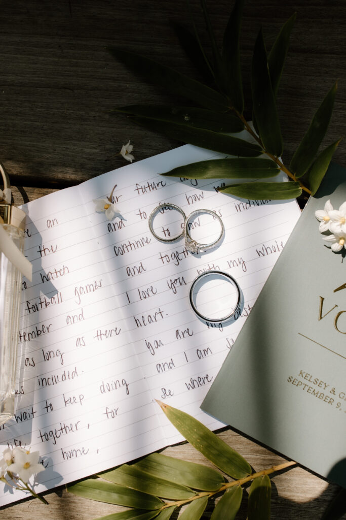 Two wedding rings rest on an open letter and a vow book. Green leaves and white flowers decorate the scene.