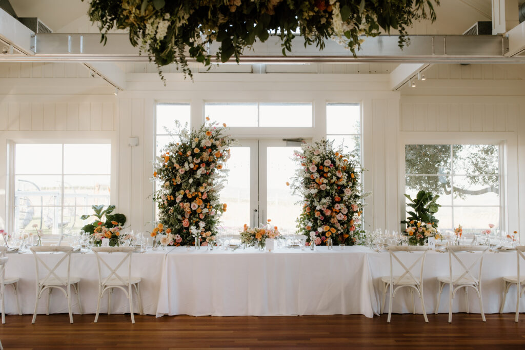 Long dining table with white tablecloth, set for an event. Floral arrangements with pink and orange flowers flank the windows, and greenery hangs from the ceiling.