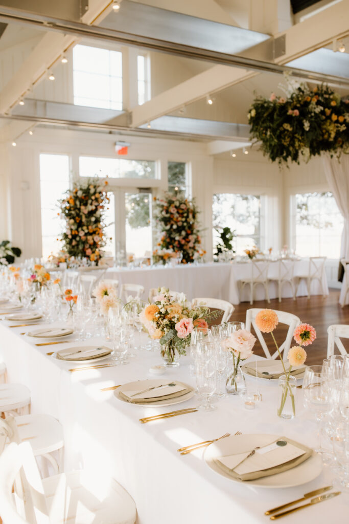 Elegant dining setup in a bright venue with long tables, floral centerpieces, and place settings. Large floral arrangements in the background, and a reflective ceiling above.