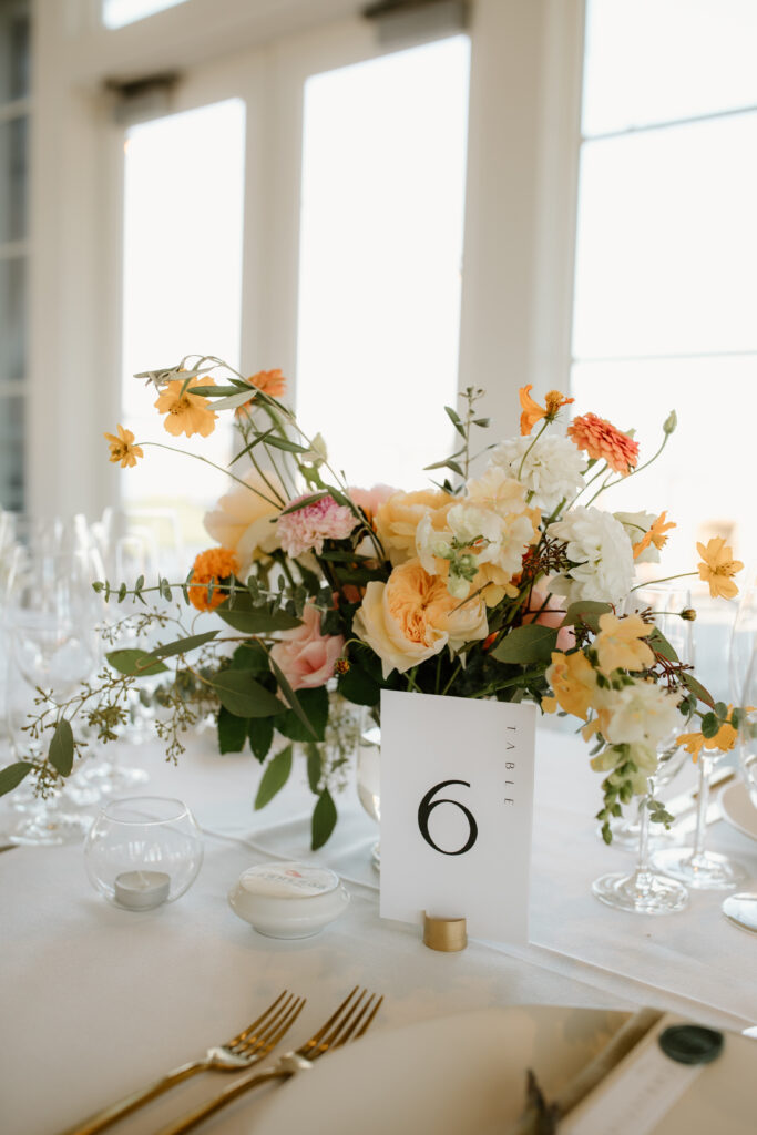 A floral centerpiece with various flowers sits on a table beside a card labeled "Table 6" surrounded by glassware and tableware.