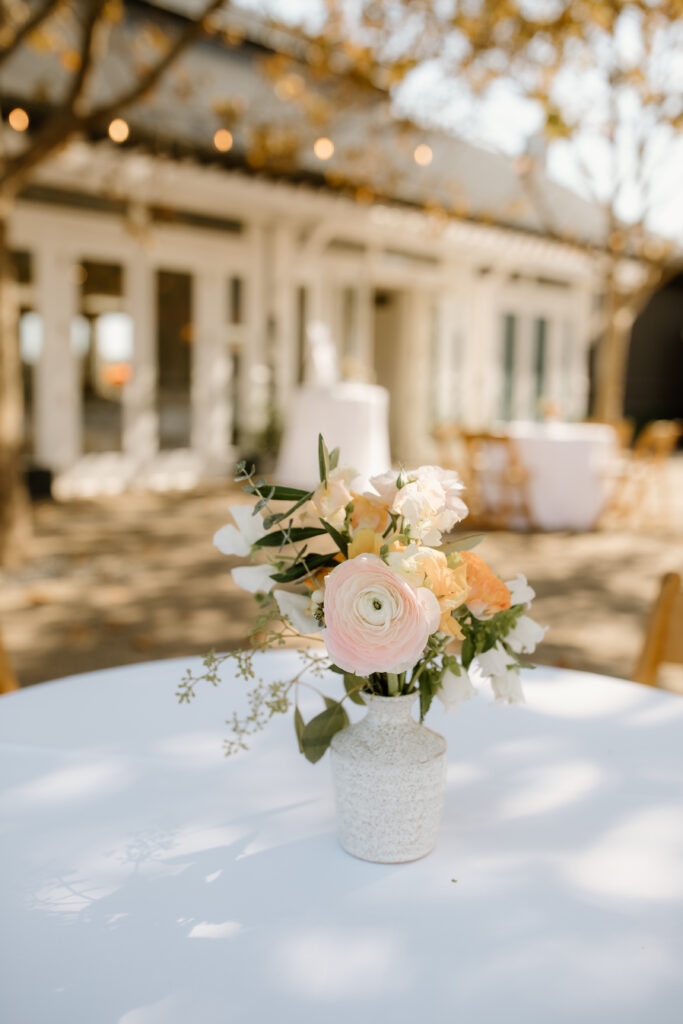 A small floral arrangement in a white vase sits on a round table outdoors, with a blurred background of a building and trees.