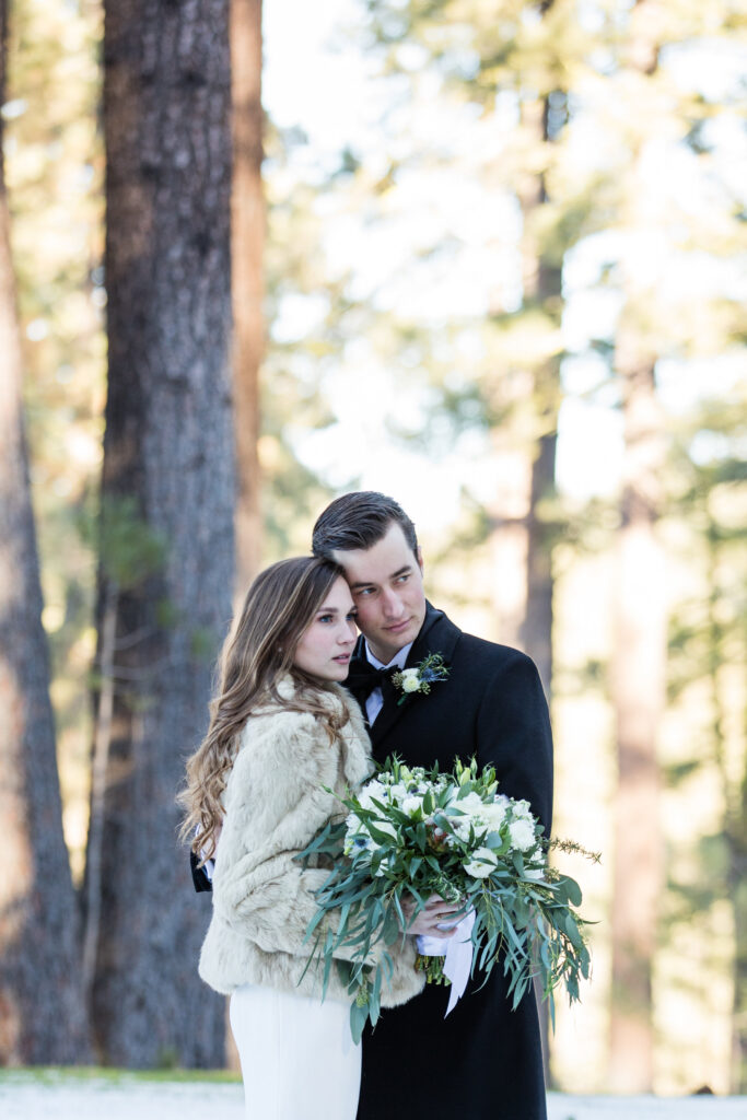 A couple embraces in a forest setting. The woman wears a fur jacket and holds a bouquet of white flowers, while the man is in a dark coat with a boutonniere. Pine trees are visible in the background.