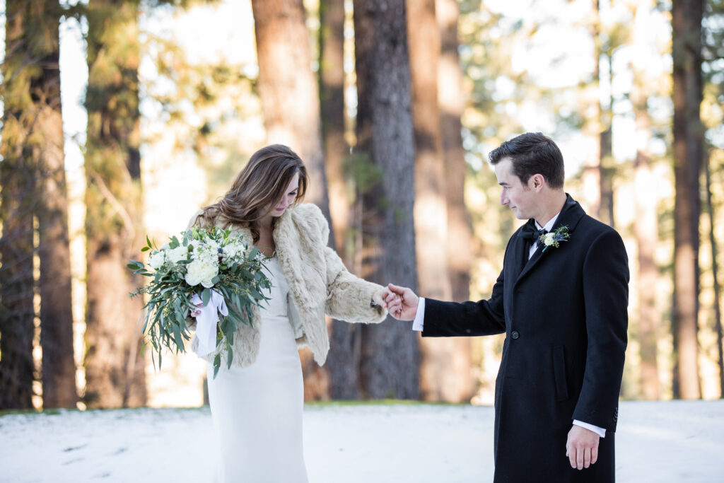 A couple stands in a snowy forest. The woman, in a white dress and fur jacket, holds a bouquet. The man, in a black coat, gently holds her hand. Tall trees are in the background.
