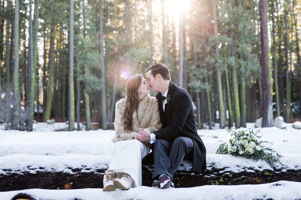 A couple sits on a log in a snowy forest, dressed warmly and holding hands, with a bouquet beside them and sunlight filtering through the trees.
