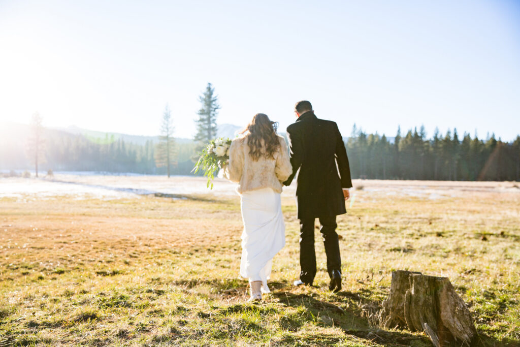 A couple in formal attire walks hand in hand across a grassy field, surrounded by trees, on a sunny day. The woman holds a bouquet.