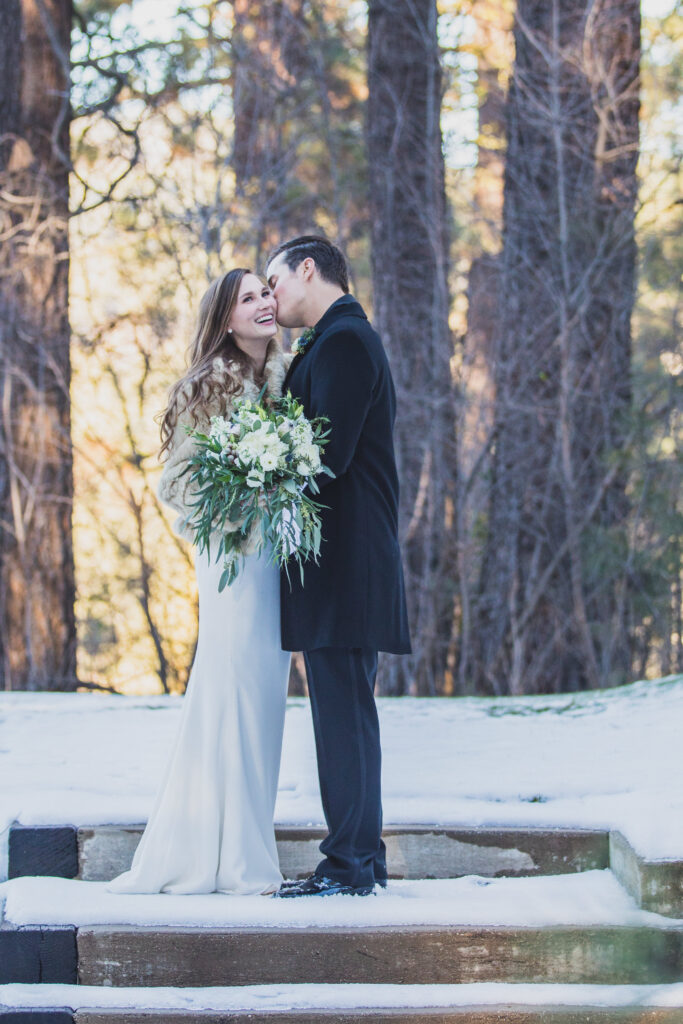 A bride and groom stand smiling on snowy steps surrounded by tall trees. The bride holds a bouquet of white flowers.