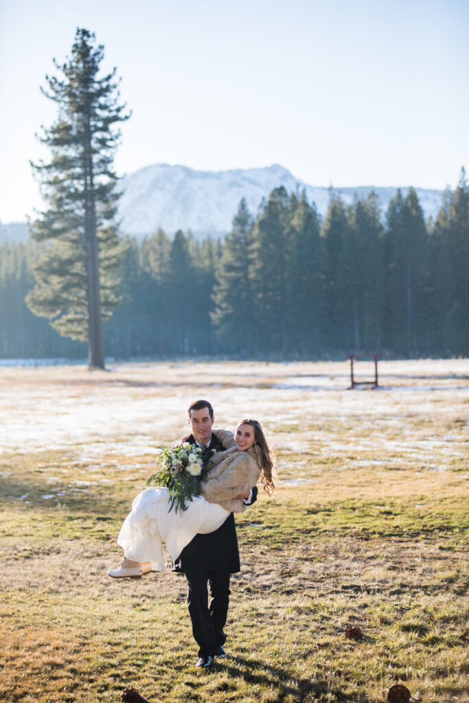 A person in formal attire carries another, dressed in a wedding gown and holding a bouquet, across a grassy field with trees and a mountain in the background.