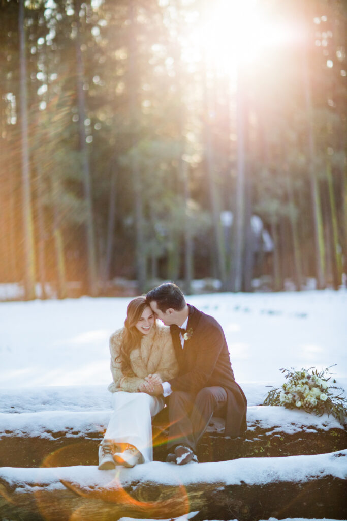 A couple sits closely on a snow-covered log in a sunlit forest, surrounded by tall trees. A bouquet rests beside them.