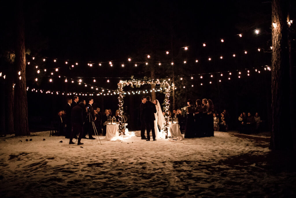 A nighttime outdoor wedding scene with a bride and groom under a flower-adorned arch, surrounded by guests. String lights hang above, illuminating the snowy ground.