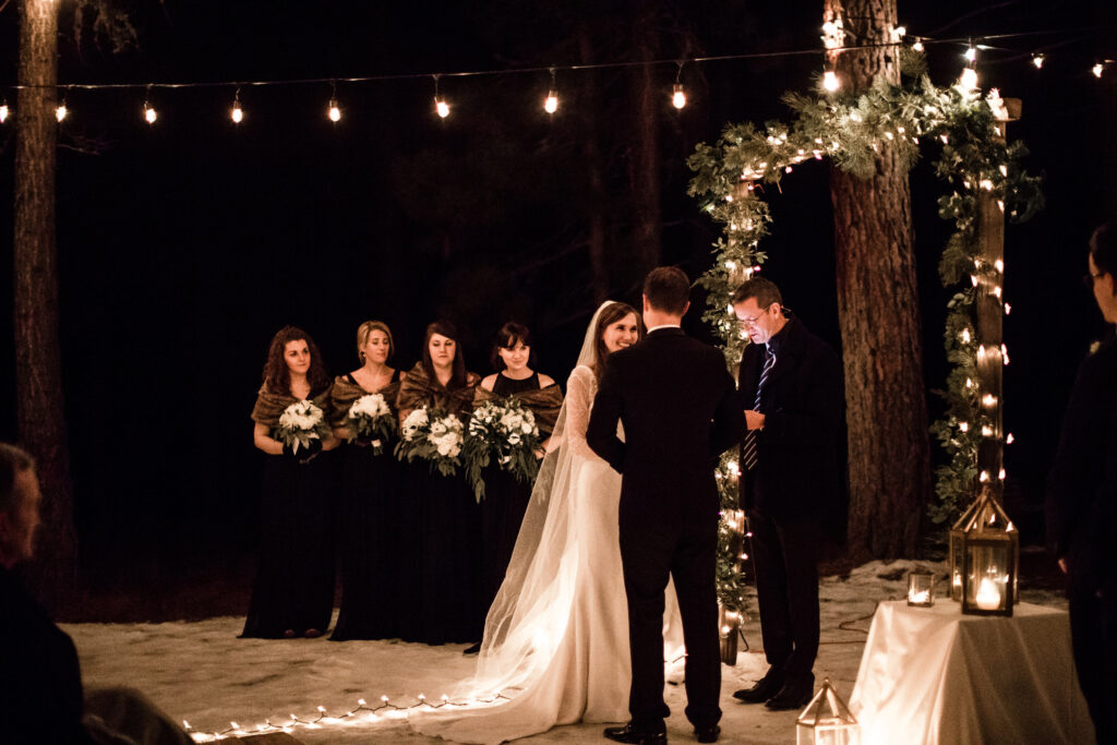 A bride and groom stand at an outdoor nighttime wedding ceremony. Bridesmaids hold bouquets nearby. Strings of lights illuminate the scene.