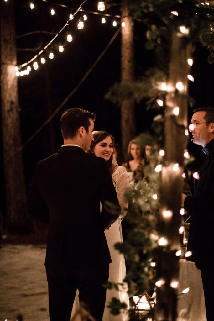 Outdoor evening wedding ceremony under string lights with a bride and groom facing each other, surrounded by trees and guests.