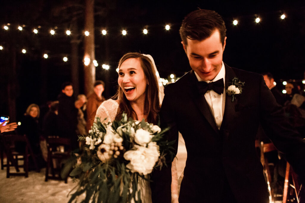 A couple in formal attire, smiling as they walk together at an outdoor evening event with string lights and guests in the background.