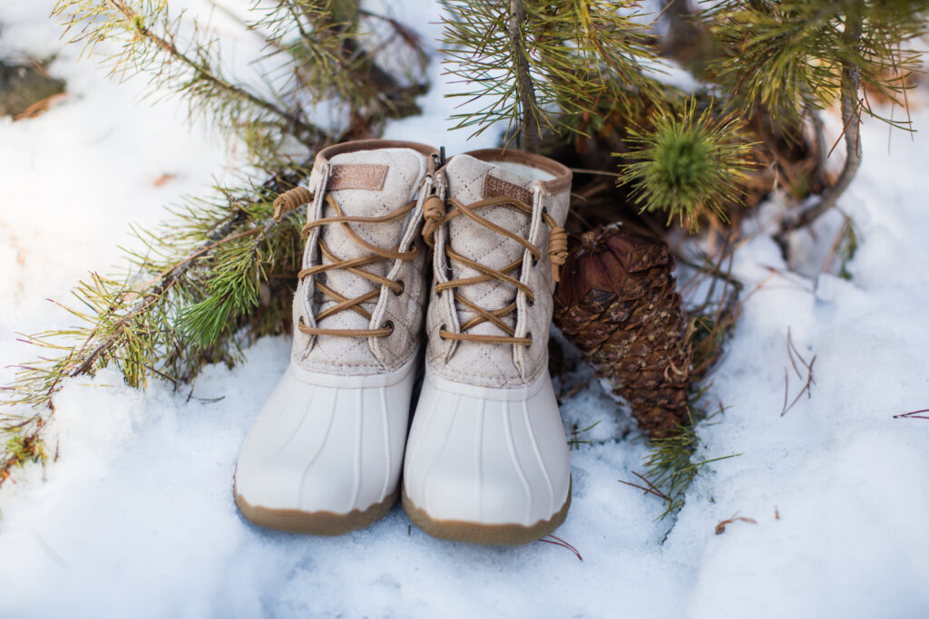 Tan lace-up boots on snow with pine branches and a pine cone nearby.