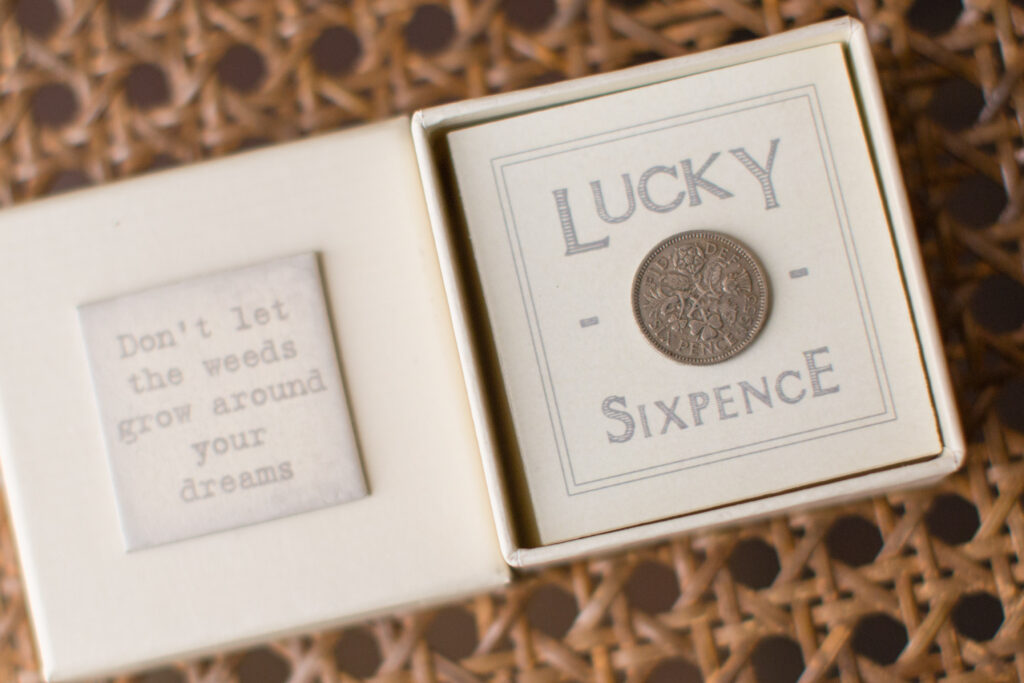 Box containing a "Lucky Sixpence" coin and a note reading "Don't let the weeds grow around your dreams" on a woven surface.