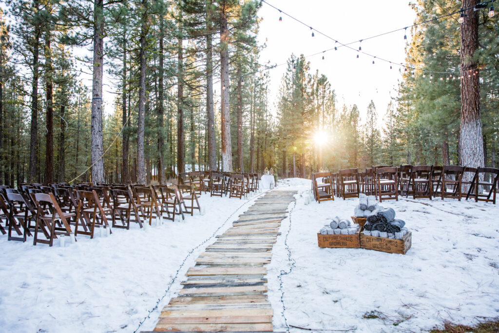 Wooden pathway through snow, flanked by wooden chairs, leading to a ceremony area in a forest. String lights hang above, and the sun shines through the trees. Firewood is stacked in crates nearby.