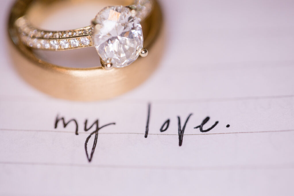 A diamond ring and a gold band rest on a lined paper with "my love" written in cursive.