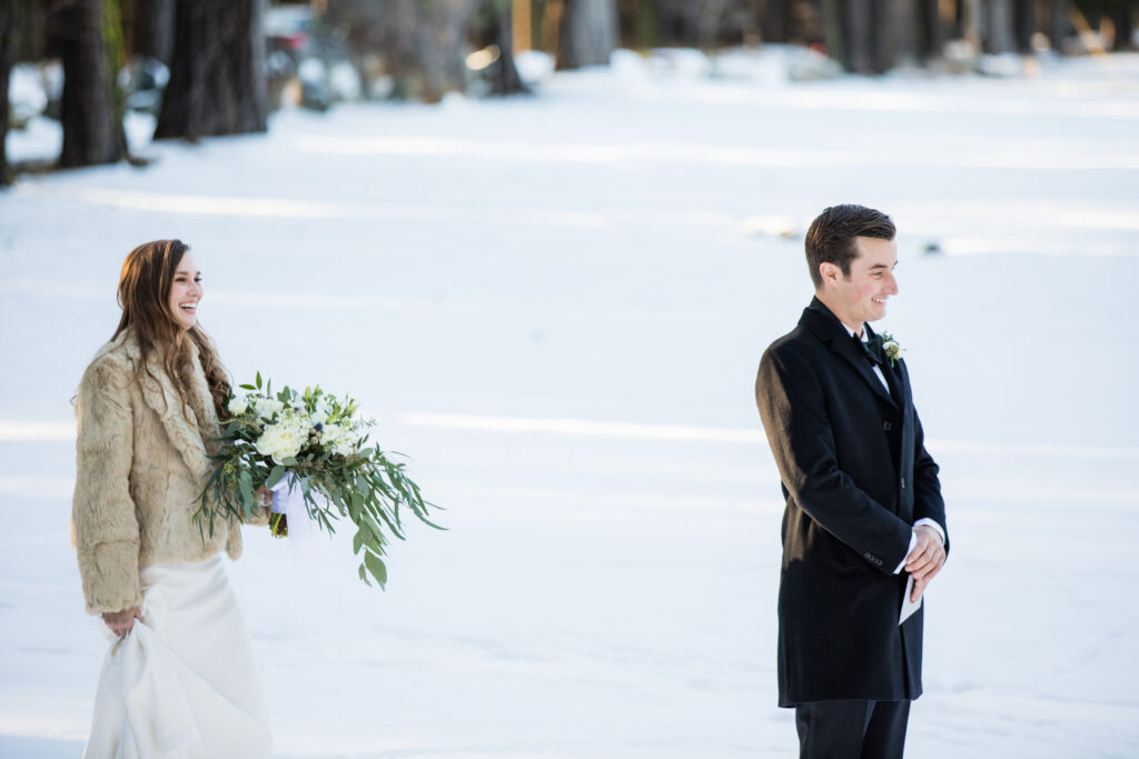 A bride in a fur coat holds a bouquet and approaches a smiling groom in a black coat on a snowy landscape.