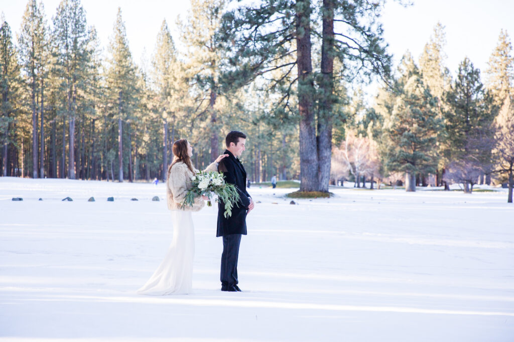 Bride in a white dress and groom in a black suit stand in a snowy landscape with tall trees. The bride holds a bouquet and approaches the groom from behind.