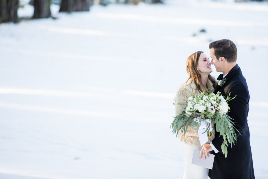 A bride and groom embrace in a snowy outdoor setting. The bride holds a large bouquet of white flowers and greenery.