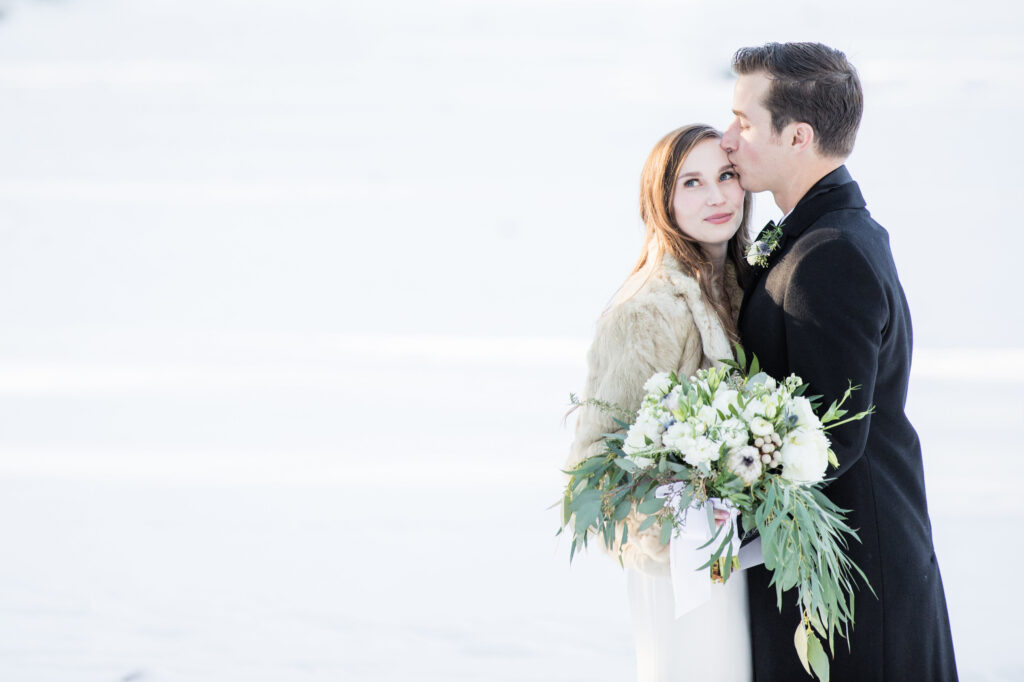 A groom in a black coat kisses the forehead of a bride wearing a fur shawl. She holds a bouquet of white and green flowers.